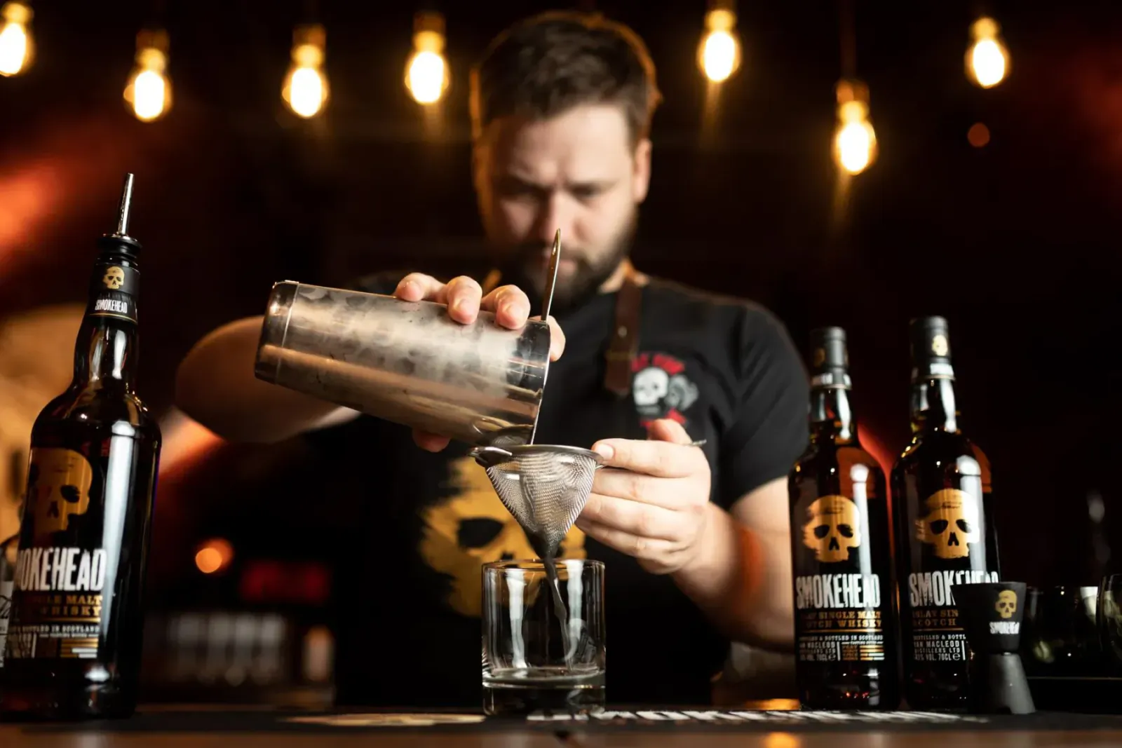 Bartender pouring Smokehead whisky cocktail from shaker into glass amid Glengoyne, Rosebank, Tamdhu bottles in dimly lit bar