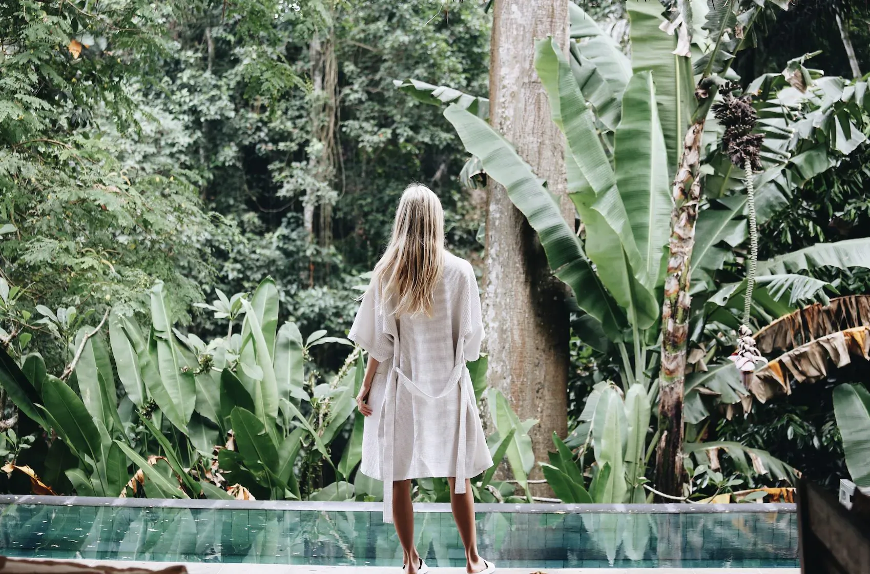 Blonde woman in white dress stands barefoot on infinity pool edge, overlooking lush Bali jungle.