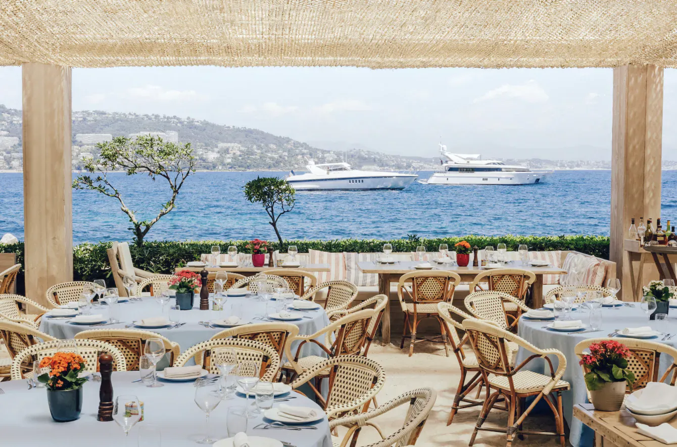 Outdoor restaurant on French Riviera with wicker tables, flowers, and yachts on blue sea beyond.
