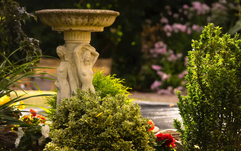 Stone pedestal birdbath with two embracing nude female statues in lush garden with pink flowers and greenery at Karma Salford Hall.