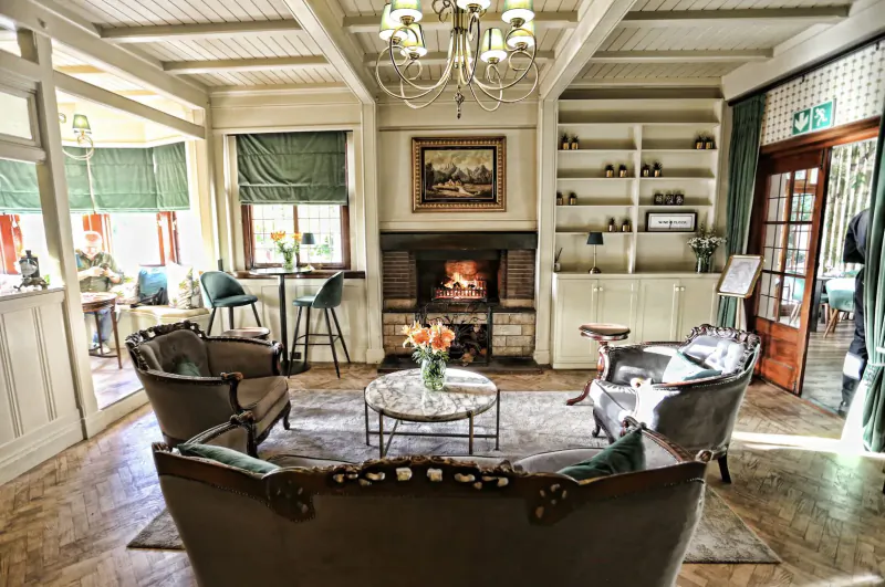 Cozy lounge at De Tafel with ornate chandelier, lit fireplace, armchairs around coffee table with flowers, bookshelves, and large windows.
