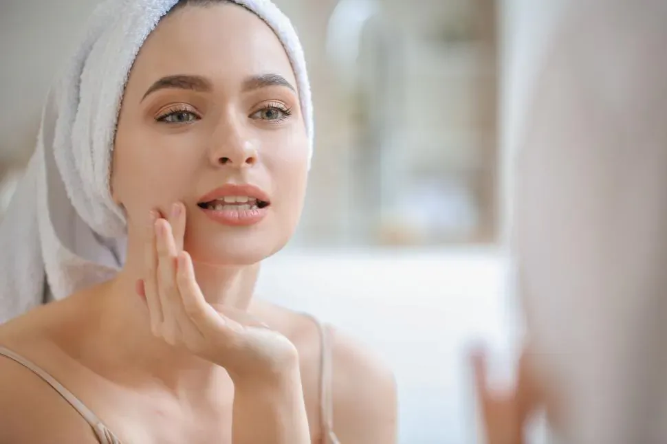 Close-up of woman with white towel on head touching her cheek, gazing in bathroom mirror