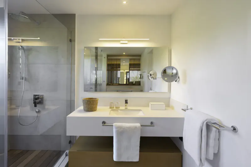 Modern bathroom in Villa Diamond, Mauritius: white vanity with towels, woven basket, mirror, frosted glass shower.