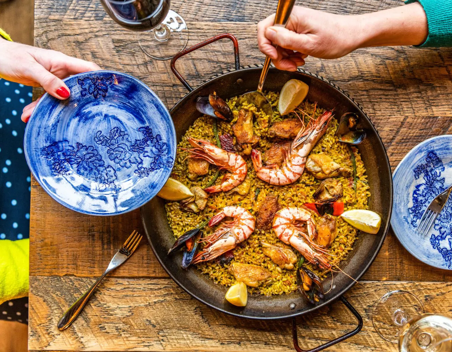 Overhead view of paella with shrimp and mussels in pan, lemon wedges, served with blue plates, chopsticks, wine on rustic wood table