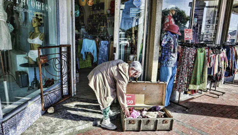 Elderly woman in beige coat bends over open trunk of clothes outside Kalk Bay clothing shop window