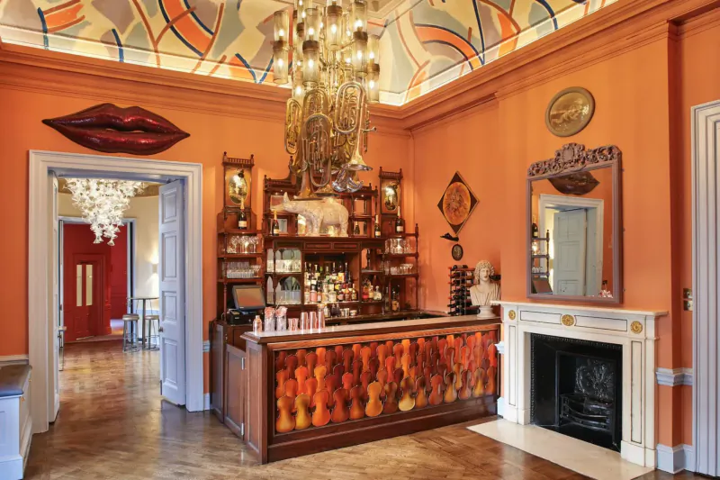 Elegant Home House bar with orange violin wall, giant lips art, chandelier, and ornate ceiling in London members club.