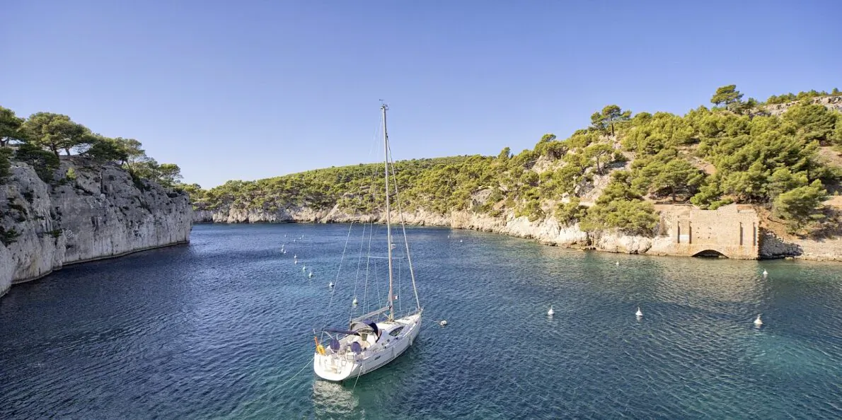 White sailboat anchored in turquoise cove surrounded by rocky cliffs and pine trees, luxury sailing destination.