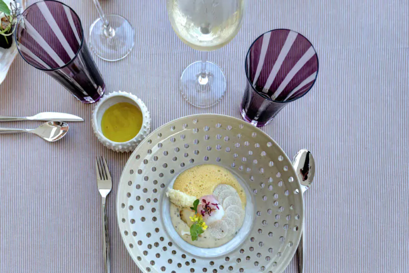 Elegant Michelin-style dish of creamy yellow custard with pink garnish and herbs in perforated bowl, flanked by wine glasses and olive oil.