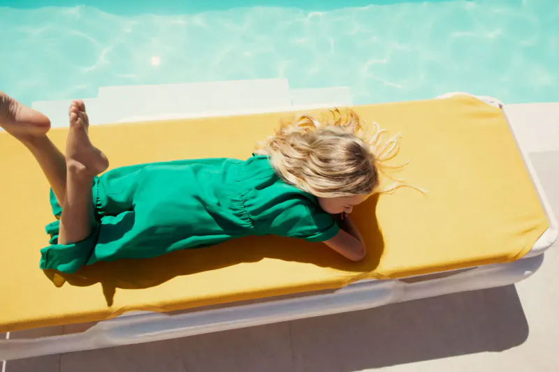 Blonde girl in green dress lying on yellow lounge chair by turquoise pool