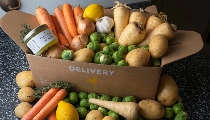 Open brown box labeled 'DELIVERY' overflowing with fresh carrots, potatoes, parsnips, onions, Brussels sprouts, lemons, and thyme.