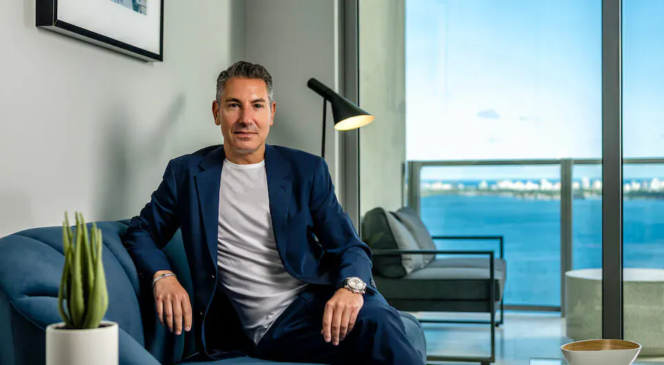 Todd Krim in navy suit seated on blue sofa in luxury high-rise living room with ocean view