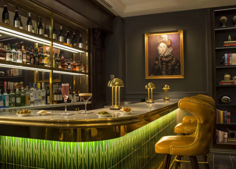 Elegant bar at The Guardsman hotel with gold stools, lit bottles, cocktails, and portrait painting.