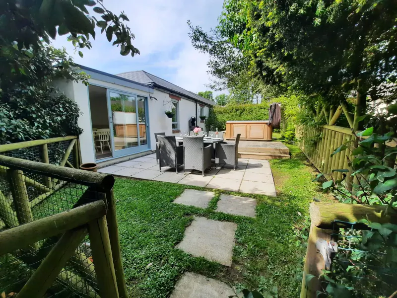 Idyllic garden patio at Broomhill Manor with outdoor dining table, hot tub, and lush greenery under blue sky