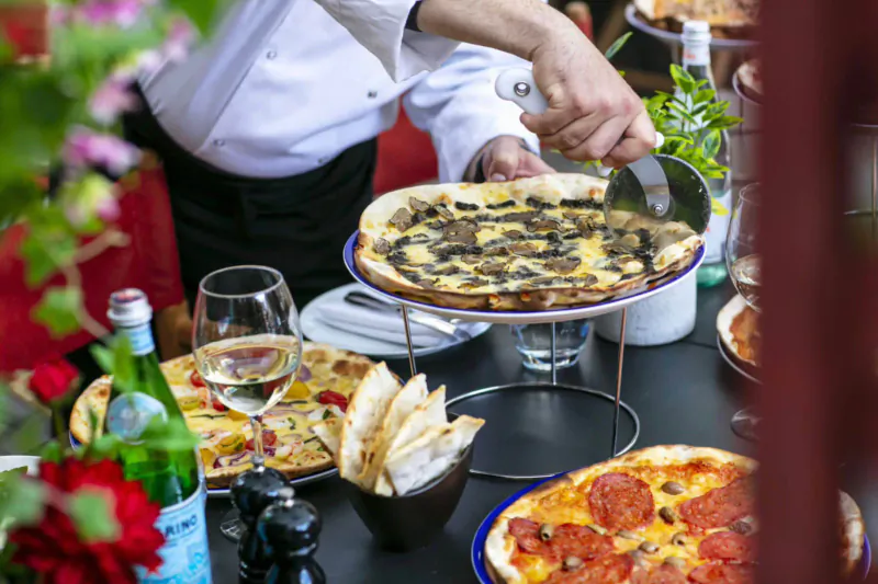 Chef in white uniform serving mushroom pizza with spatula at table with wine, salads, and other pizzas at Crazy Pizza Marylebone.