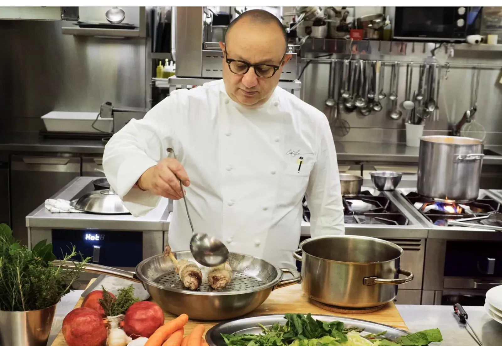 Bald chef in white uniform ladles meat from wok in professional kitchen with veggies nearby