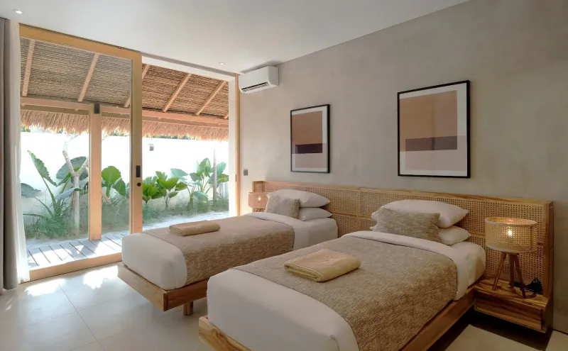 Cozy bedroom in beachfront villa with two wooden twin beds, beige linens, lamps, abstract art, and garden view through sliding doors.