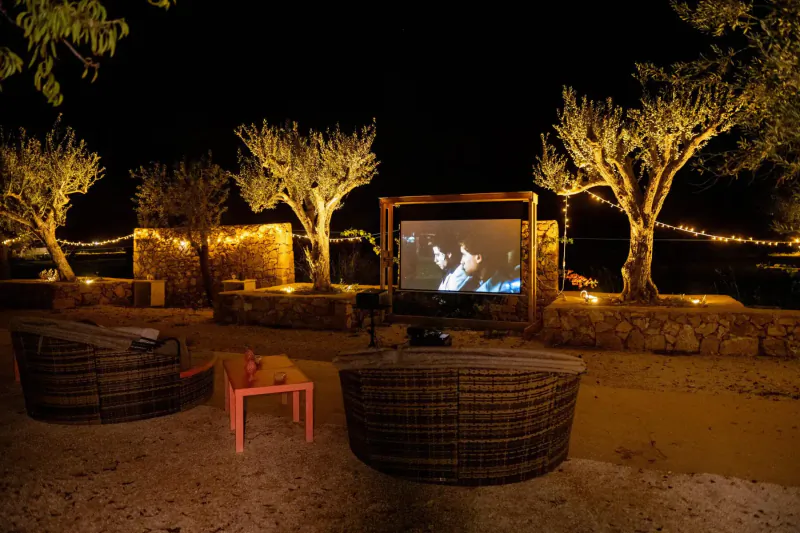 Outdoor cinema at Zahir Country House: large screen under wooden canopy, rattan chairs, olive trees, stone walls, night fairy lights in Sicily.