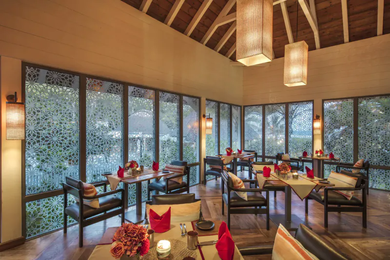 Elegant resort dining room with wooden ceiling, lanterns, red napkins on tables, and tropical garden views through windows.