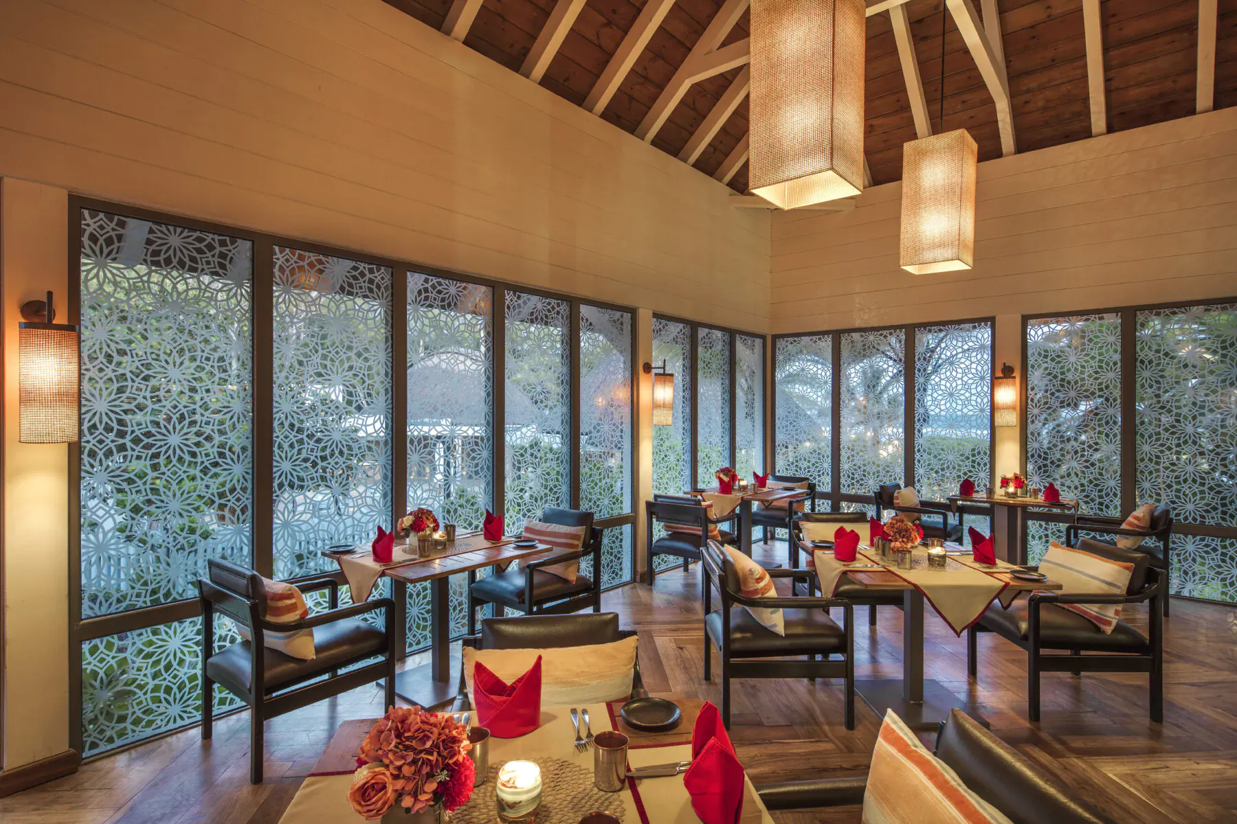 Elegant resort dining room with wooden ceiling, lanterns, red napkins on tables, and tropical garden views through windows.