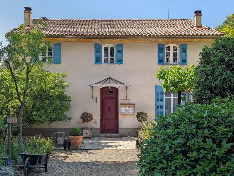 Lou Calen Boutique Hotel facade in Provence: ochre house with blue shutters, red door, potted plants, gravel path, trees.