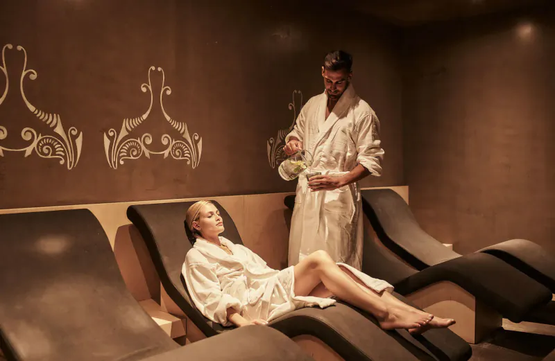 Man in white robe pours drink for blonde woman reclining on lounge chair in spa lounge at Elysium Boutique Hotel.