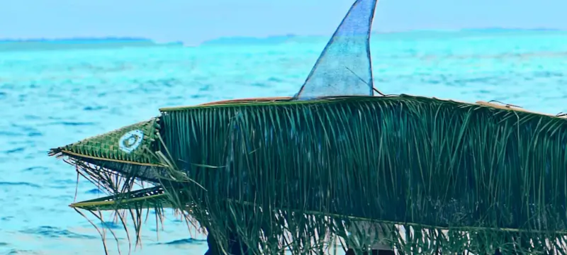 Large woven palm frond shark sculpture with open mouth and blue fin on turquoise ocean shore at beach resort