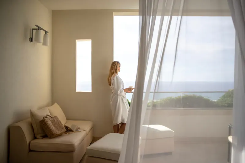 Blonde woman in white robe stands by window holding cup, gazing at sea view through sheer curtains in luxury hotel room