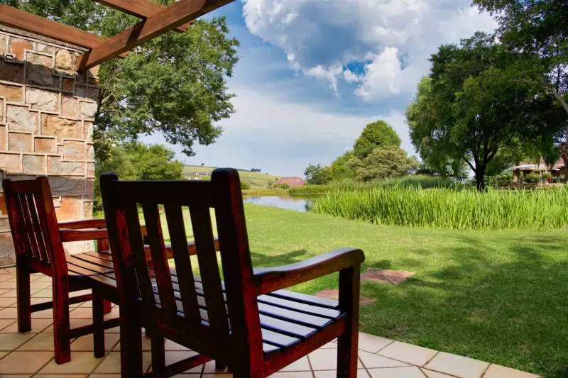 Two wooden chairs on a patio overlooking lush green lawn, river, trees, and hills under partly cloudy sky.