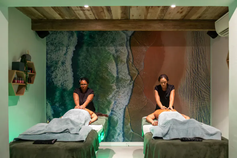 Two masseuses giving massages to clients on beds in Coastal Calm Oyster Suites spa with ocean mural backdrop.