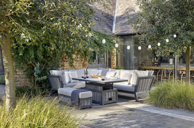 Modern grey Bramblecrest L-shaped outdoor sofa set with coffee table on garden patio amid olive trees and string lights.