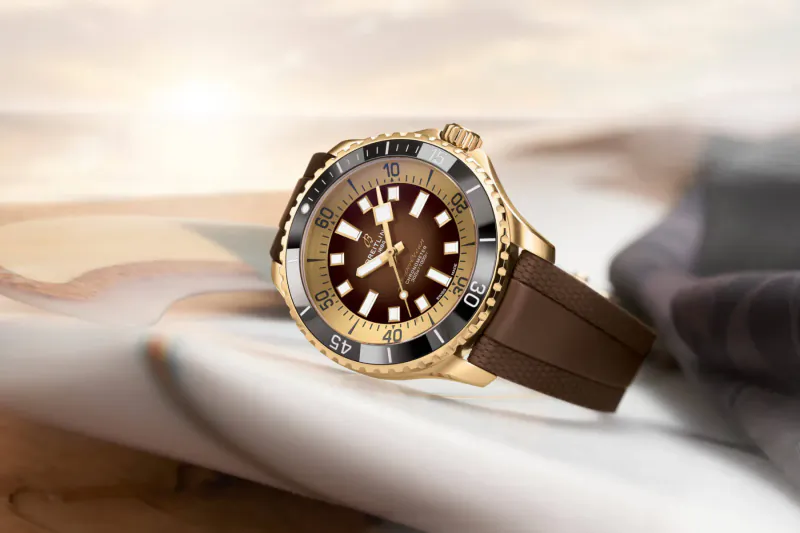Close-up of gold and brown Breitling Superocean dive watch with brown rubber strap on a surface, warm sunset glow.