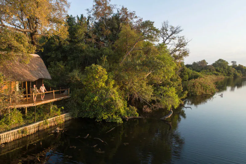 Aerial view of Xugana Island Lodge, thatched cabins on wooden deck by river, two people standing, surrounded by lush trees at sunset.