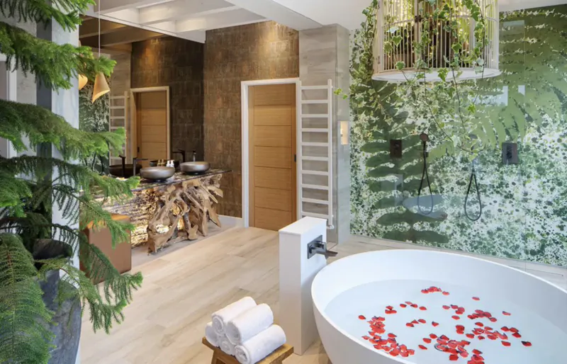 Luxurious Gilpin Hotel bathroom with white bathtub filled with red rose petals, wooden table, green fern wall, trees, and towels.