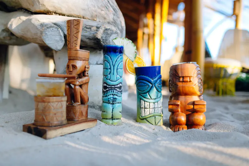 Carved tiki mugs on sand: brown tikis flank blue cocktail with lime and pineapple at tropical beach club.