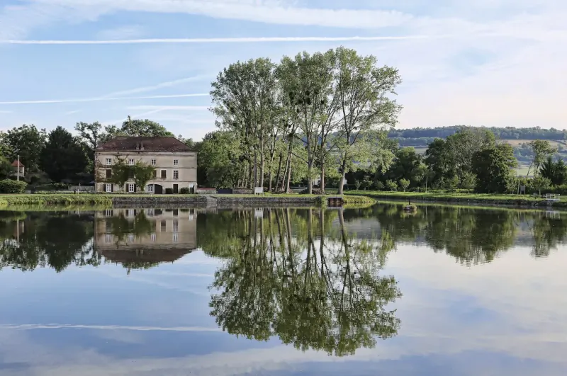 Beige chateau with red roof reflected in calm pond, surrounded by green trees and hills in Burgundy wine country.