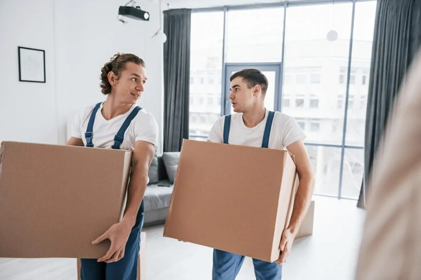 Two smiling movers in white overalls carry cardboard boxes in a modern living room with large windows.