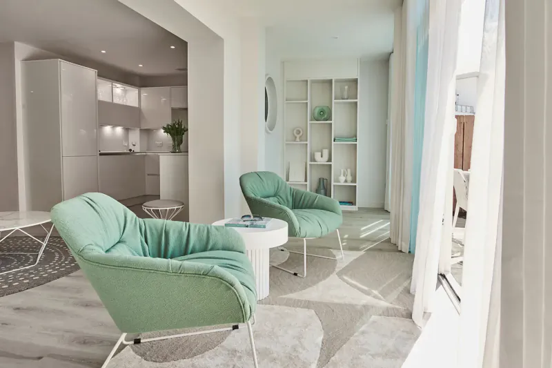 Modern luxury living room at St Moritz Hotel with two mint green armchairs, glass table, white kitchen, and large windows.
