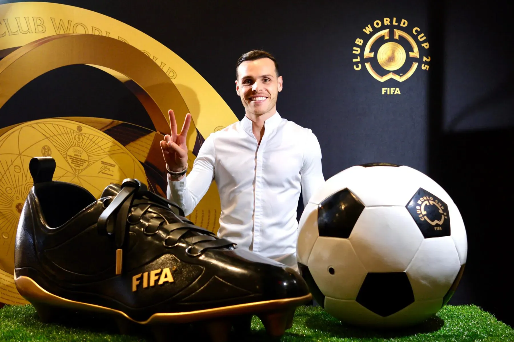 Amaury Guichon peace sign beside giant black FIFA sneaker and soccer ball on FIFA Club World Cup backdrop