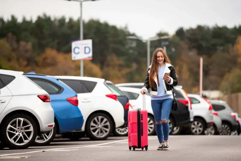 Smiling young woman in leather jacket pulling pink suitcase past cars in Gatwick Airport CD parking area