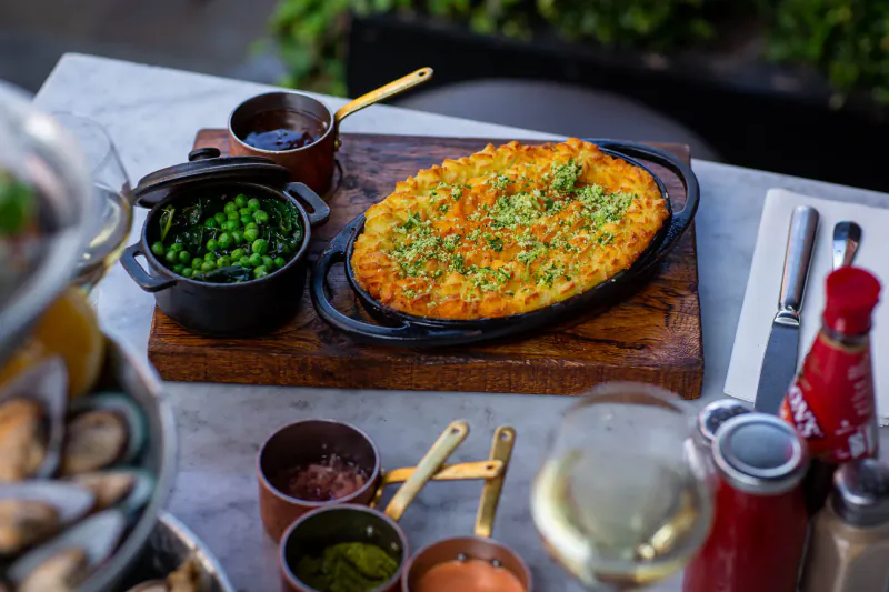 Oval cheesy fish pie topped with peas, served with peas, sauces, wine, ketchup, and condiments on a wooden board at Lobster Pot Booth.