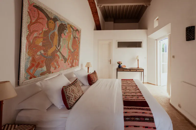 Luxurious bedroom in Moroccan villa with white bed, colorful textiles, abstract wall art, and beamed ceiling.