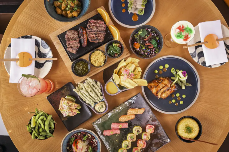 Overhead view of Sushi Samba fusion platter: sushi rolls, grilled meats, edamame, salads, and cocktails on wooden table