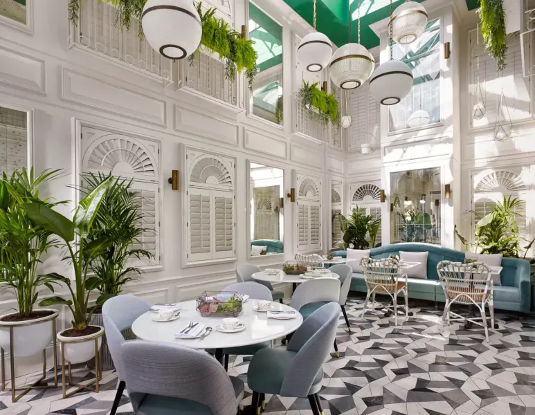 Elegant atrium dining room with white walls, green ceiling, hanging plants, blue velvet chairs, round tables, checkered floor.