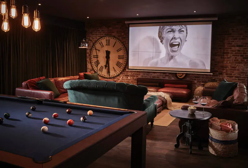 Cozy lounge in Harper Norfolk with pool table, plush sofas, large clock on brick wall, and projected woman's portrait.