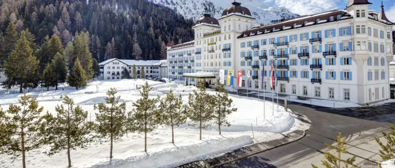 Grand Hotel des Bains Kempinski St. Moritz, elegant white alpine hotel with flags, surrounded by snowy pines and mountains.