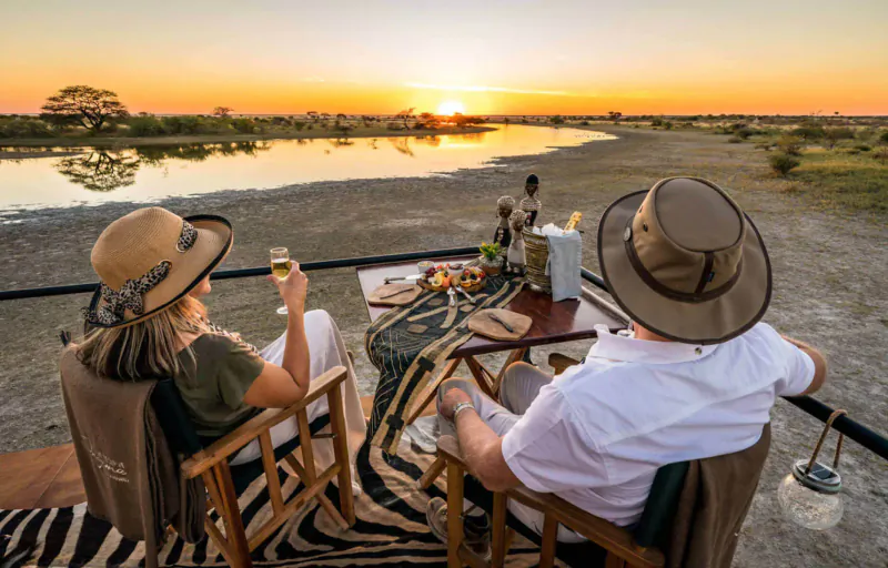 Couple in safari hats toasting wine at candlelit table with food, overlooking Etosha savanna river at sunset.