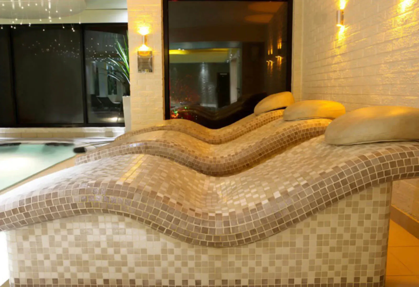 Three curved mosaic loungers beside indoor pool at Moddershall Oaks Spa, warmly lit with wall sconces.
