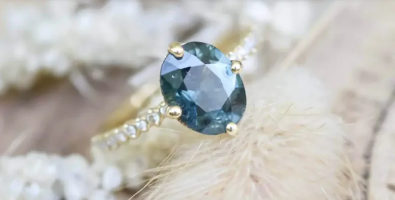 Oval teal gemstone engagement ring with diamond pavé gold band on fluffy white flowers