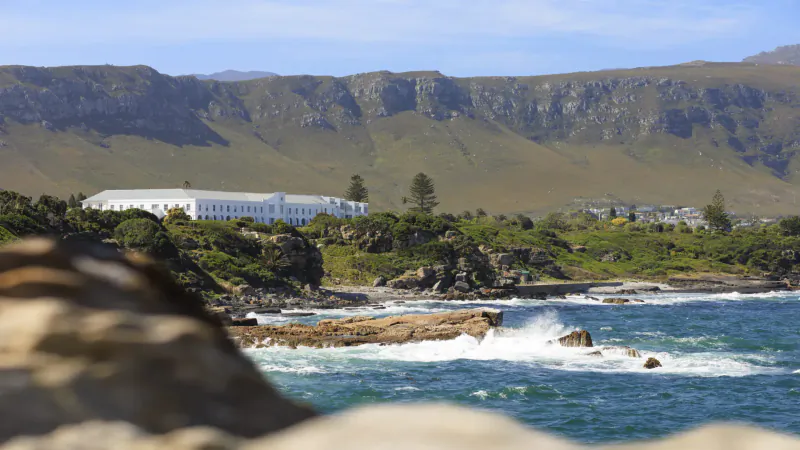 The Marine Hotel Hermanus, white building on green hills above crashing ocean waves and rocky coast, South Africa