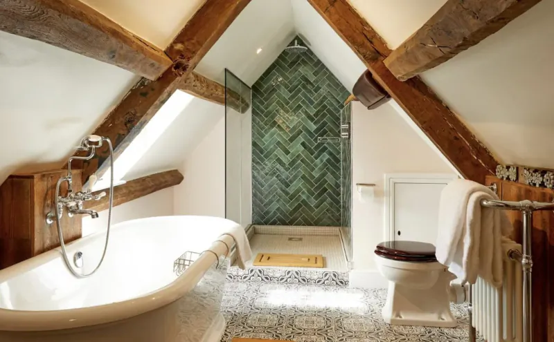 Cozy attic bathroom with exposed wooden beams, freestanding bathtub, green tiled walk-in shower, and toilet.